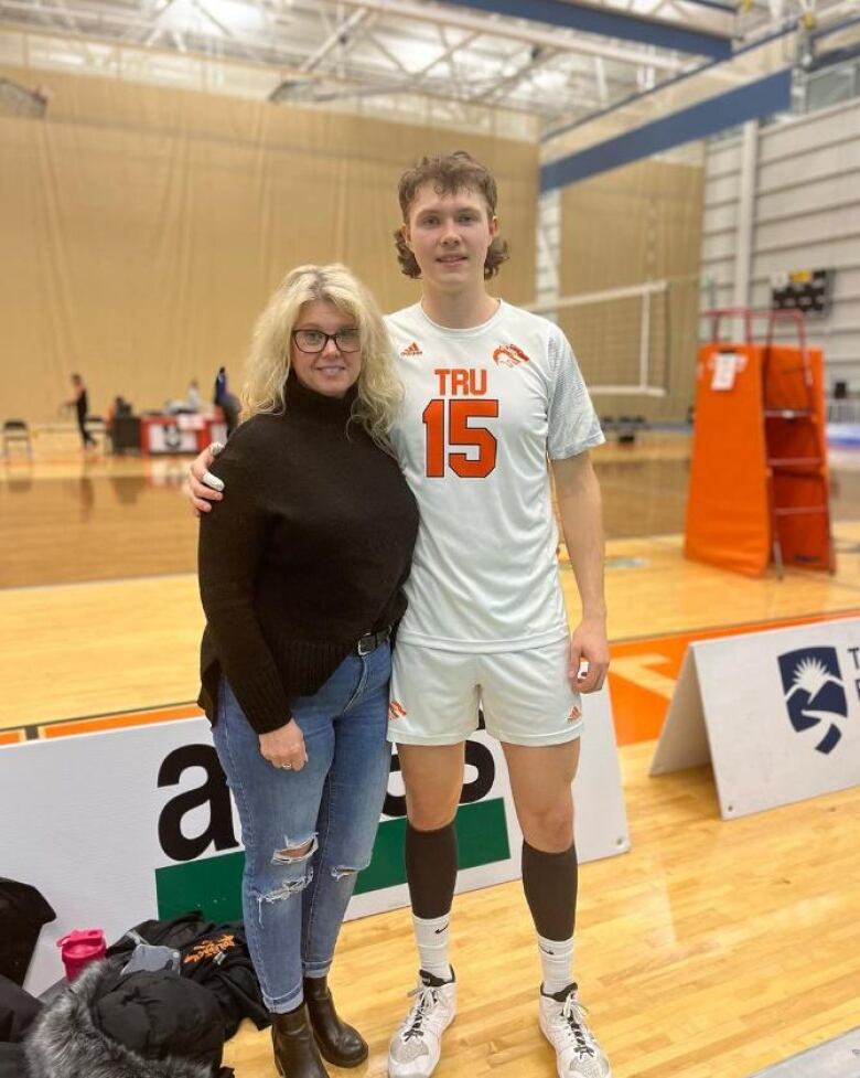 A blonde woman stands next to a tall young man in a volleyball jersey inside a large gymnasium.