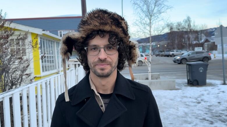 A man wearing a hat stands on a street and looks into the camera. 