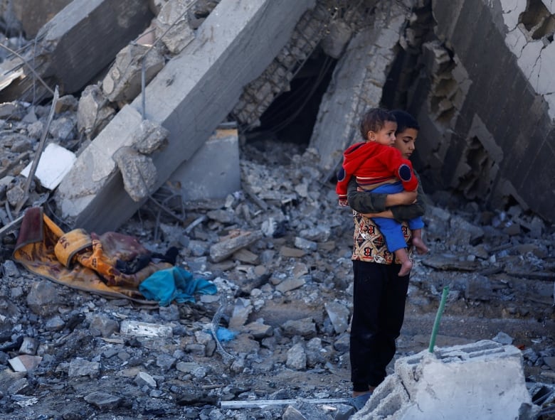 An older boy holds a small child on his shoulder as he stands amid concrete rubble and debris outdoors.