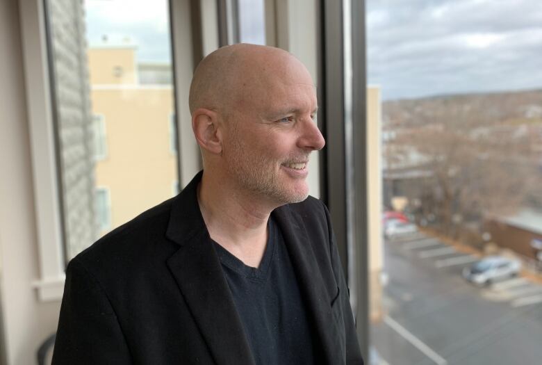 A Caucasian man wearing a black blazer looks out a second-storey window over an urban landscape. 