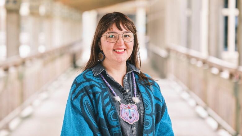 A woman wearing a blue denim shirt with a blue shawl and a pendant around her neck. She's wearing glasses and has brown hair.