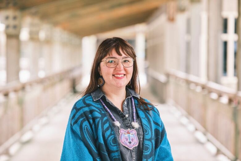 A woman wearing a blue denim shirt with a blue shawl and a pendant around her neck. She's wearing glasses and has brown hair.