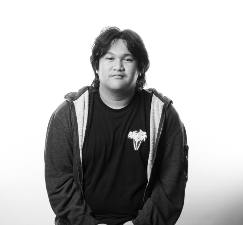 Young man with shaggy, dark, shoulder-length hair sits on a stool in this black and white portrait. He is wearing a dark, unzipped sweatshirt with a black T-shirt underneath. He is wearing grey sweat pants and his hands are clasped on his lap. 