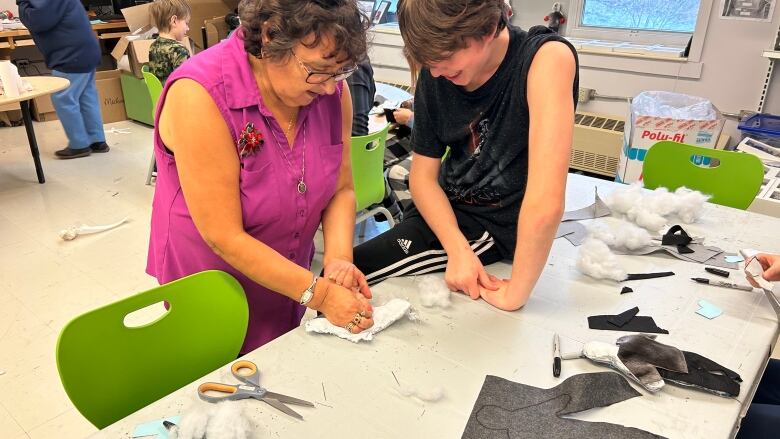 A woman standing next to a student who is sitting on a table full of crafting items. The woman is showing the boy how to sew.