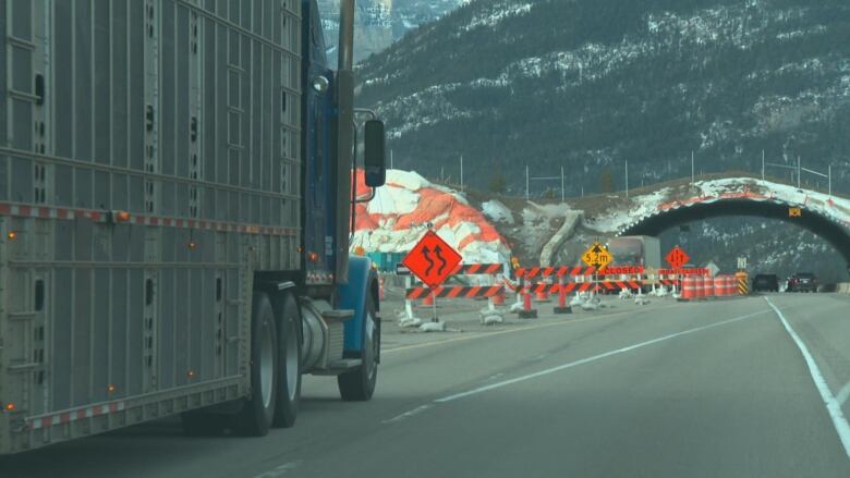 A semi drives toward an underpass with construction signs around it. 