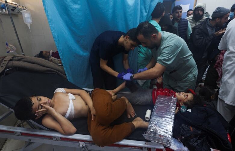 A young boy, wearing brown pants with a bandage wrapped around his chest, lies on a stretcher in a hospital room. Beside him, medical staff treat a young girl who is crying.