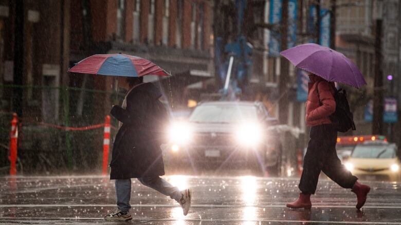 Carrying umbrellas, two pedestrians walk across a crosswalk amid heavy rain, with vehicles and their front lights on facing them. 