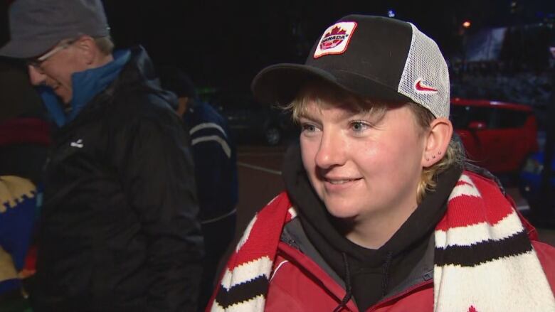 A white woman in her 30s wears a red jacket, white scarf and Team Canada hat while standing in a parking lot.