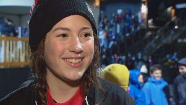 A teen girl with a long brown braid wears a Team Canada black and red toque, a red shirt and a black windbreaker while standing on the sidelines of a stadium with a crowd behind her.