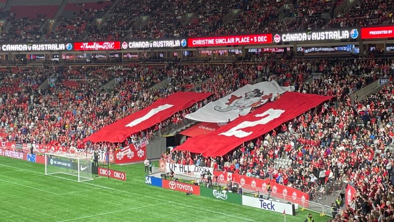 Fans hold three large banners; left banner is red with the number one, the middle banner has a women's soccer player celebrating, the right banner is red with the number two.