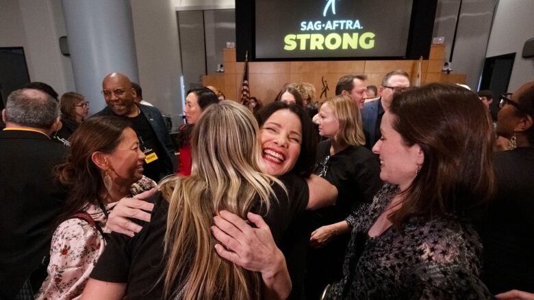 A dark-haired woman who face is to the camera hugs another woman in a croweded room. In the background, is a sign that reads SAF-AFTRA strong.