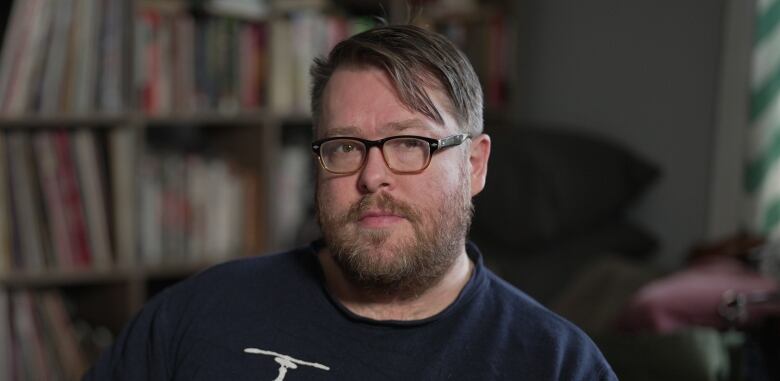 A man with glasses and a beard, and wearing a blue t-shirt, looks thoughtfully at the camera.