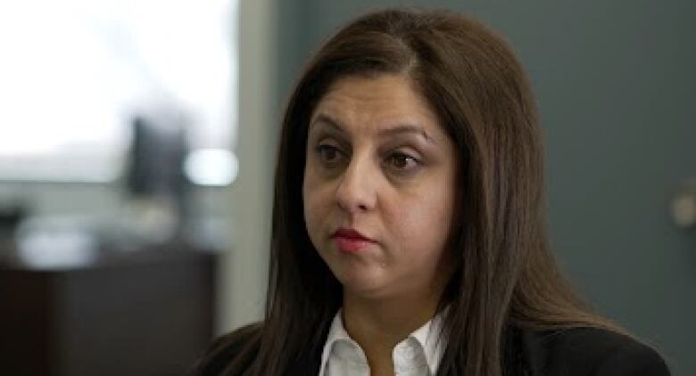 A woman with long dark hair, wearing a dark blazer, is seen sitting in an office with a serious expression on her face.