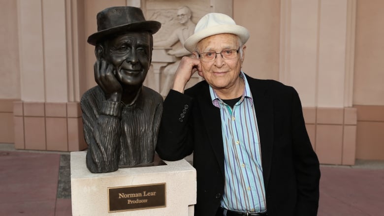 A man mimics a statue of himself leaning his head against his right hand. Statue and man both wear the same hat. The statue is on a pedestal with a plaque that says, 