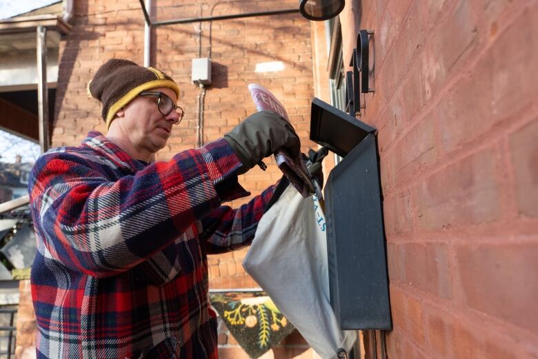 A man wearing flannel, glasses and a fall clothing delivers a newspaper to a letterbox on a front stoop.