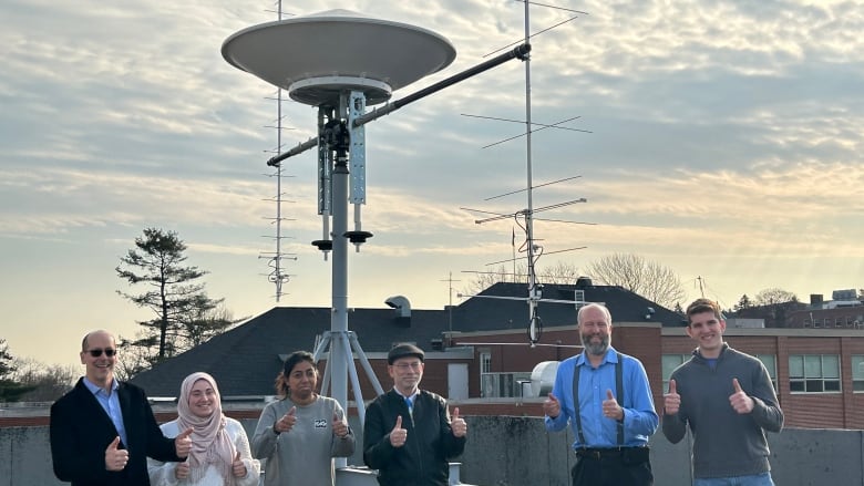 A team of six posing with their thumbs up, in front of a tall metal structure on a building's roof.