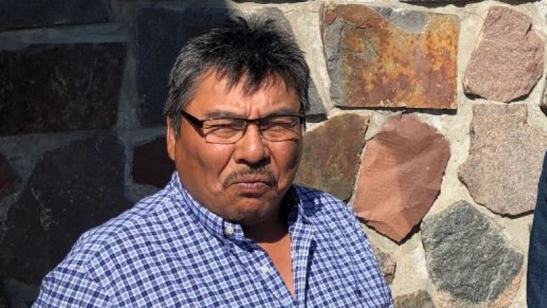 A man in a checkered shirt squinting into a camera. It appears to be sunny outside, and he's standing in front of a brill wall. 