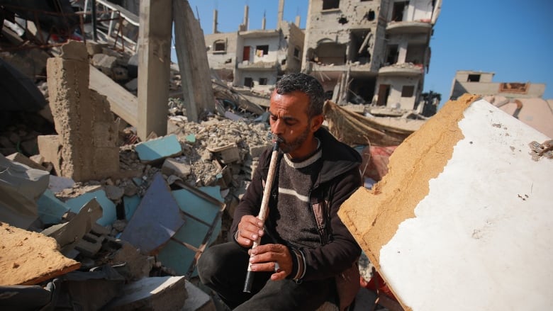 Man plays a homemade flute amid building rubble.