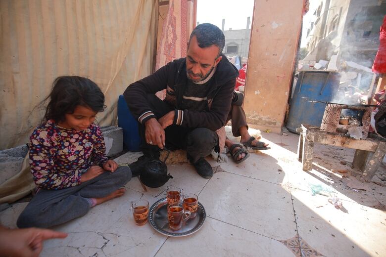 A man pours tea for a small child amid building rubble.