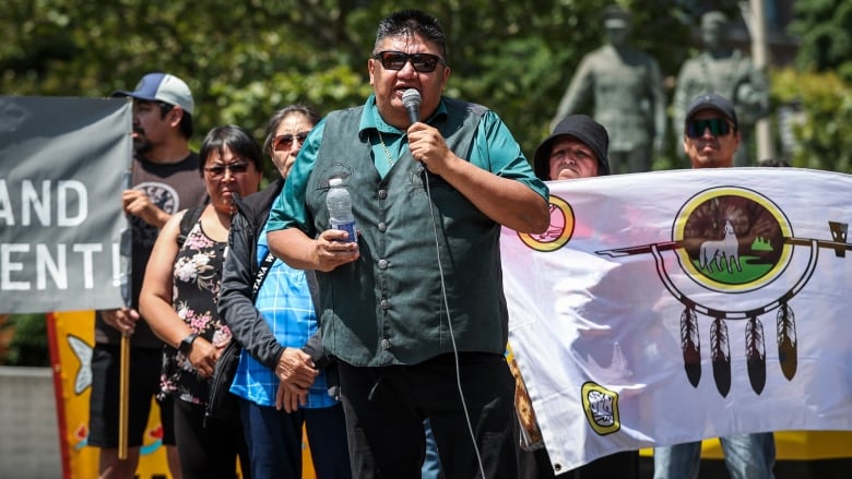 A person stands and speaks into a microphone. A crowd of people stand behind him, holding a large flag.