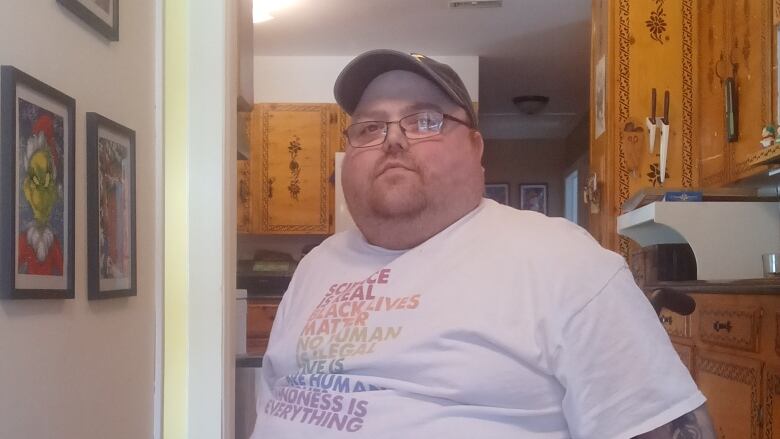 A man in a white T-shirt and black ball cap sits in a wheelchair in his kitchen.