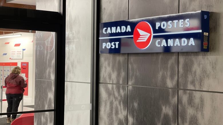 A Canada Post sign is illuminated against a faux seal skin wall, as a woman stands in line inside the new post office.