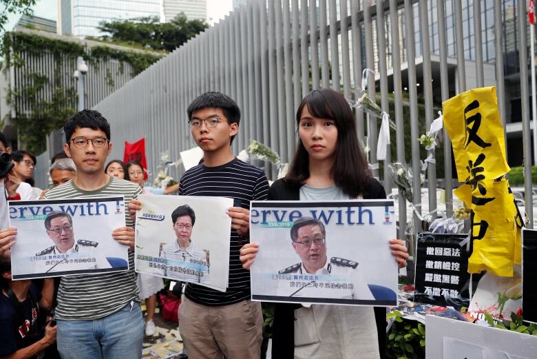 Two bespectacled Asian men and an Asian woman hold up pictures of other individuals at a demonstration outdoors.