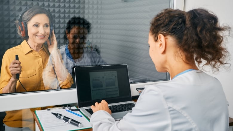 A woman wearing headphones sits across the glass from another woman in front of a laptop. 