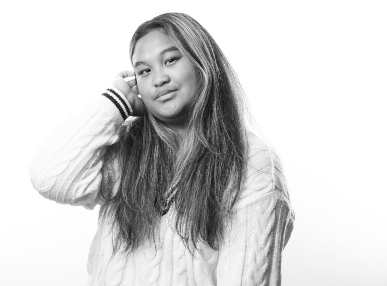 Black and white portrait of a young women with long, straight dark hair with lighter strands. She is wearing a white, cable knit sweater and dark pants. Her right hand is reaching up to push her hair behind her ear. 