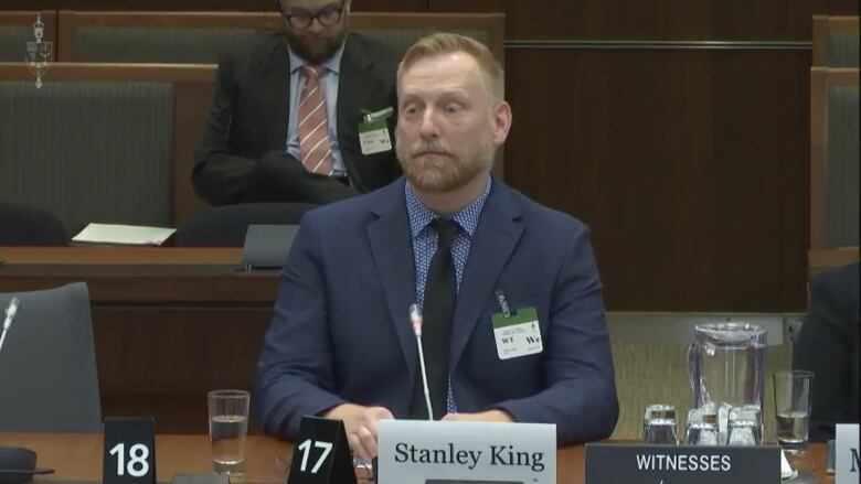 A man in a blue serge suits speaks into a microphone with a desk sign saying Stanley King in front of him