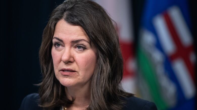 A woman's mouth is nearly closed as she speaks, with Alberta and Canadian flags behind her.