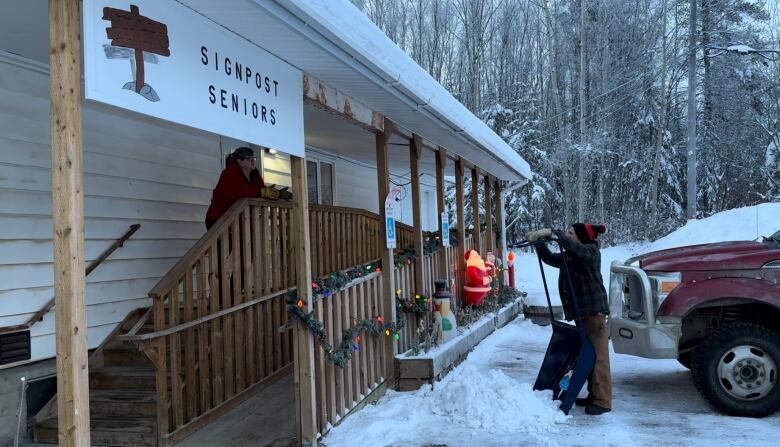 A man with a snow shovel in a parking lot talks to a woman standing on the porch of a building.