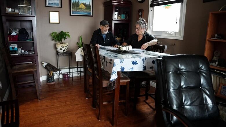 Two people sit at a dining room table with a stack of papers in front of them. 