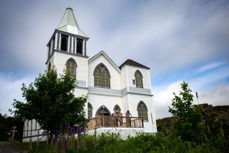 A white clapboard church with a large belltower stands in front of trees and greenery.