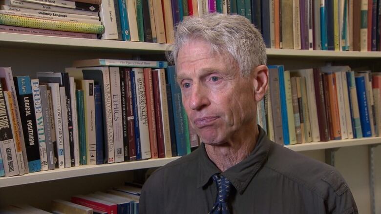 A man with short gray hair in a dark shirt and dark blue tie stands next to a book shelf.