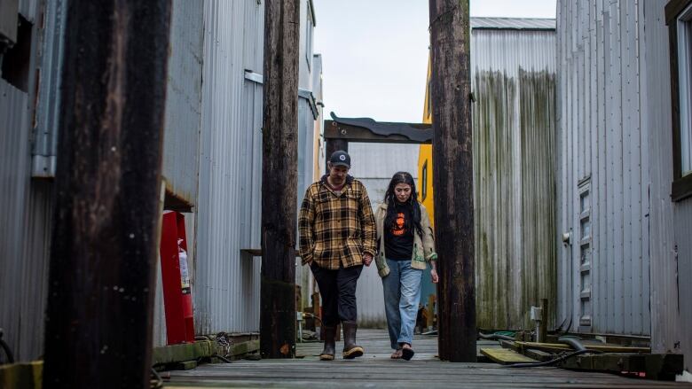 A man and a woman walk along a wooden dock, between log poles and metal-clad sheds.