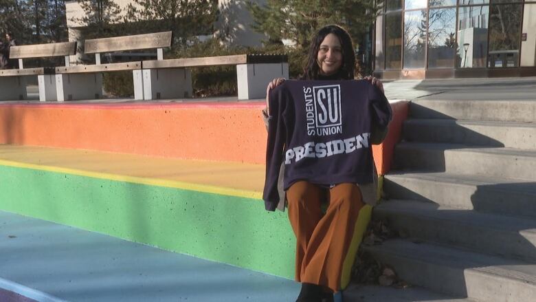 A woman sits on rainbow stairs holding a sweatshirt that reads 'Students' Union President.' 