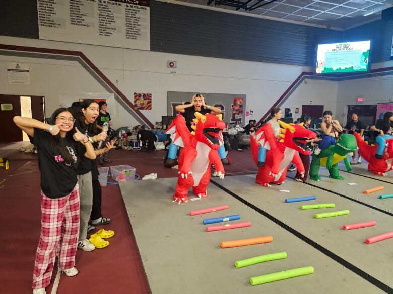 A group of students playing games. Some of them are wearing inflatible dinosaur costumes. There are foam rollers organized in a ladder formation in front of them. 