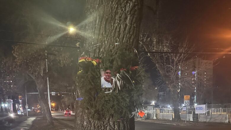 A wreath with a photo in the middle of it hangs from a tree next to a sidewalk. 