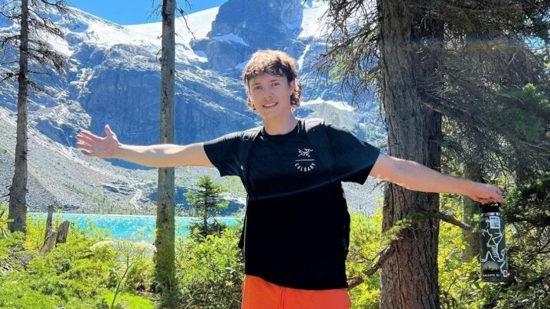 A 22-year old young man holds his arms wide on a hiking trail with a lake and snowy mountains in the background.