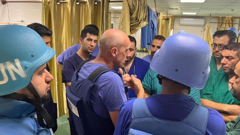 Three people in blue vests and helmets speak with people wearing hospital scrubs.