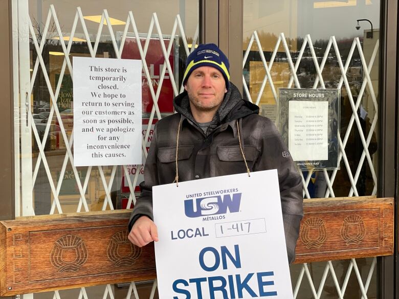 A man holding a sandwich board reading 'USW Local 1-417 ON STRIKE' stands next to a closed store door.