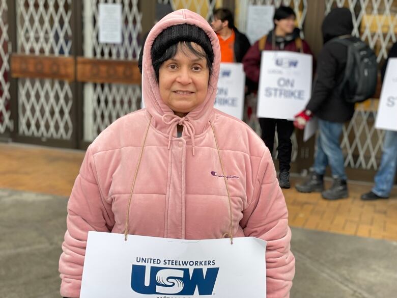 A woman in a pink puffer jacket smiles as she holds up a sign on a picket line.