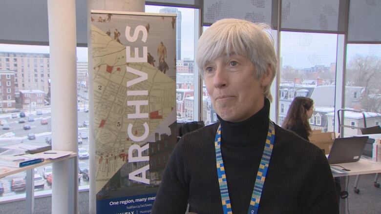 A woman with short grey hair wearing a black turtleneck speaks to the camera.