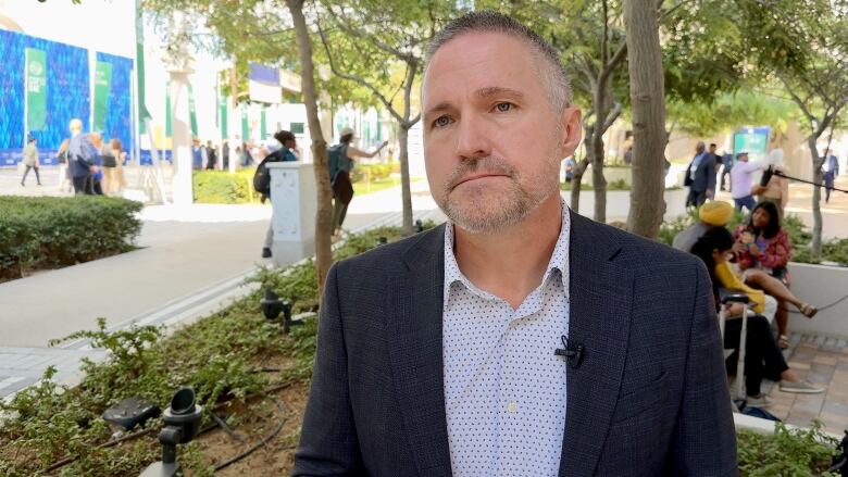 A man is interviewed outdoors at a conference venue with people walking and sitting in the background.