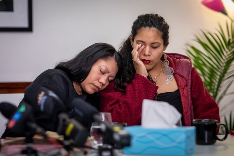 Two Indigenous women console each other as they sit in front of microphones at a news conference.