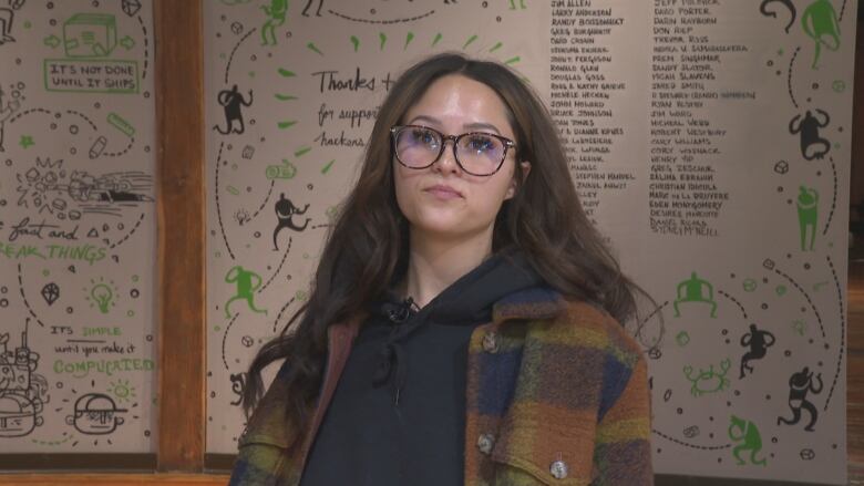 A woman with brown hair and black-rimmed glasses stands in front of a white board with doodles and writing. 