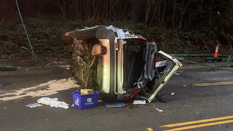 A flipped over car lies broken to pieces on a road, with a blue recycling bin near the rear wheel.