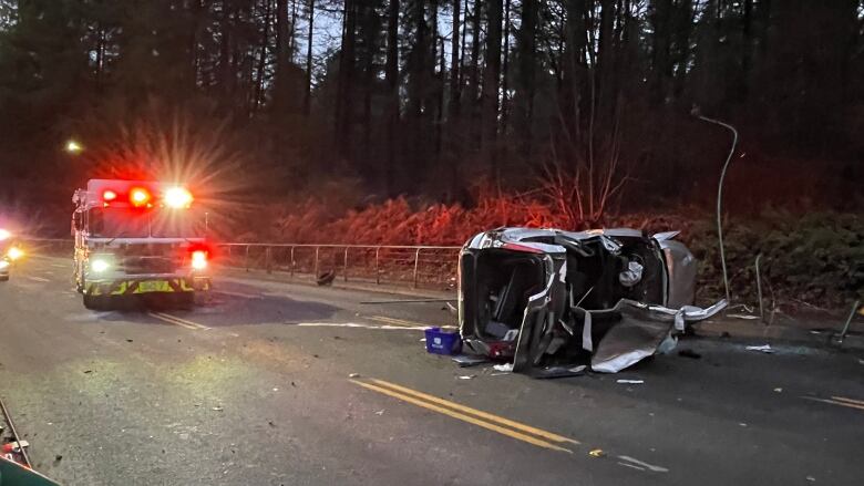 A flashing fire truck next to a destroyed and flipped over car on a road.