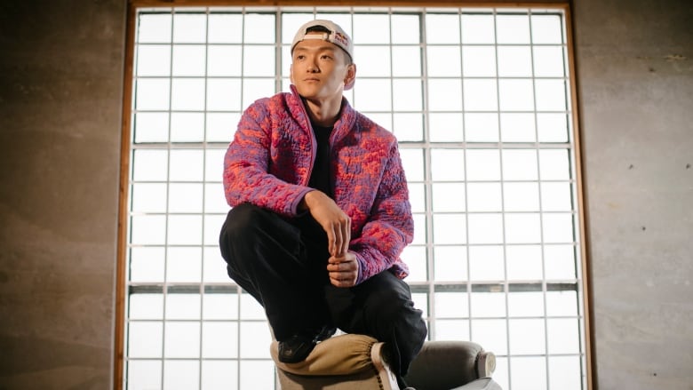 A young man wearing a baseball cap turned backwards, a colourful jacket and black pants perches on the back of an armchair.